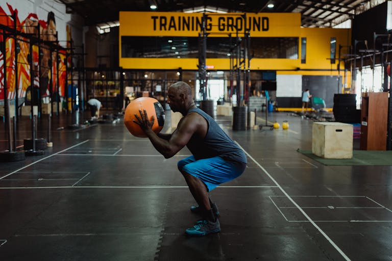 Side view of an African American man squatting with a medicine ball in an indoor gym setting, showcasing fitness and strength.