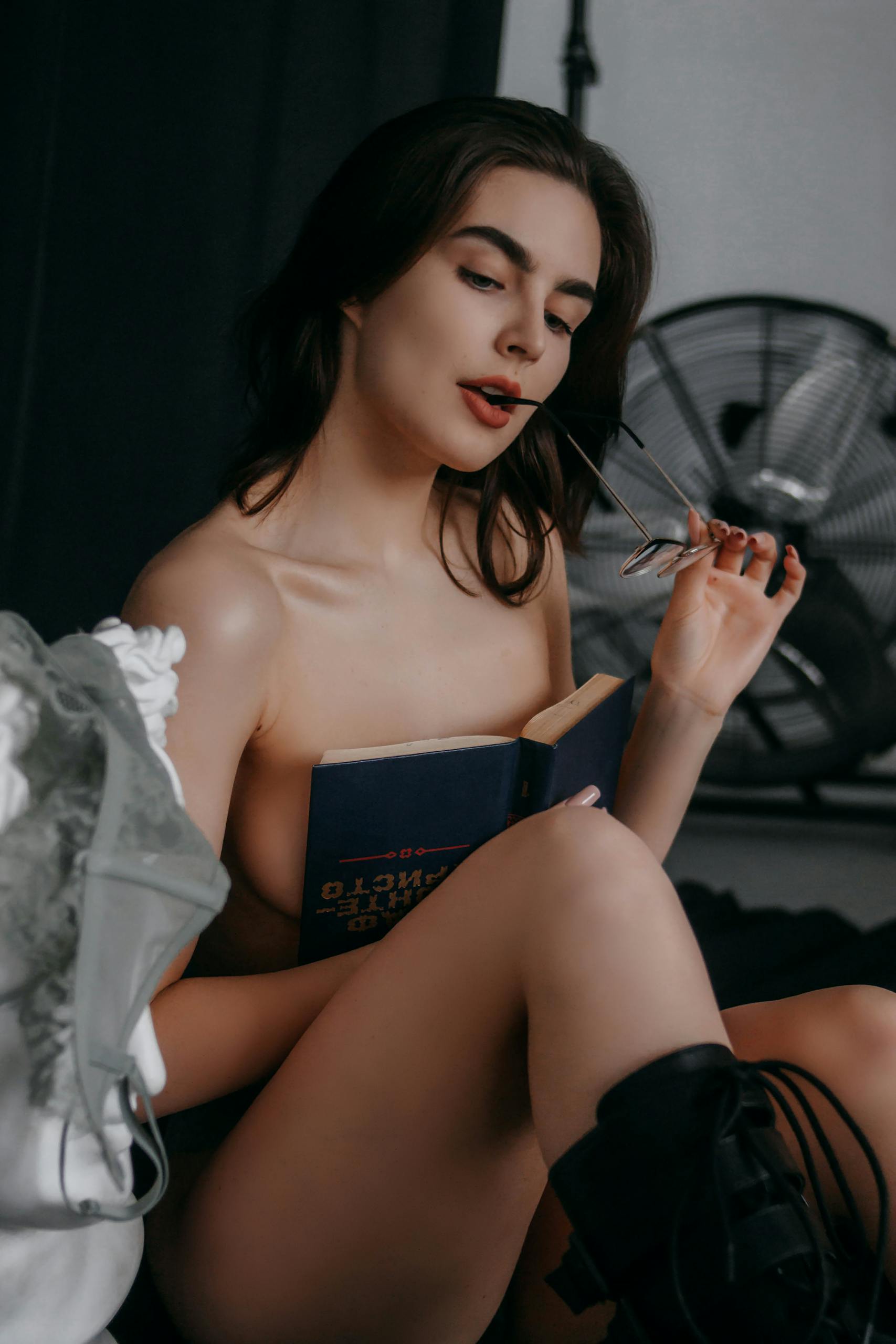 Sophisticated woman with glasses in an artistic pose, holding a book in a dimly lit studio.