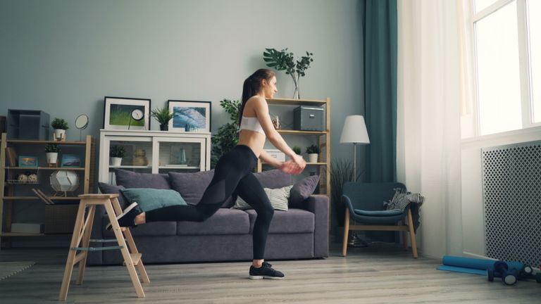 A woman in sportswear working out indoors using a chair for support, demonstrating leg exercises.