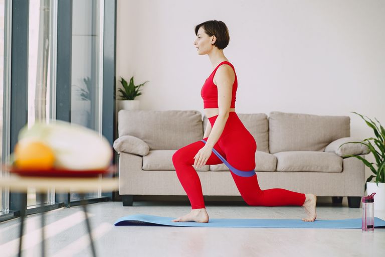 Side view of fit female in sportswear with short hair doing exercise with elastic band on blue mat near sofa in living room
