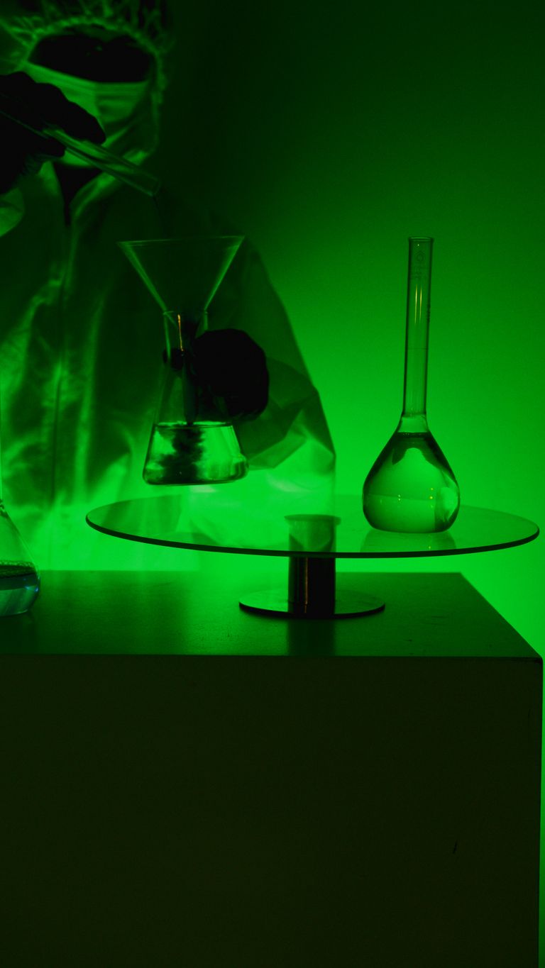 A scientist in protective gear handling glassware in a green-lit laboratory setting.
